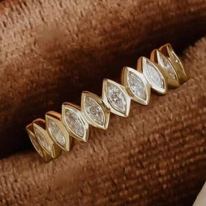 May include: A gold ring featuring a repeating pattern of marquise-cut gemstones. The ring is presented in a brown velvet jewellery box. The clear gemstones sparkle, and the gold band has a polished finish. The ring is likely for a special occasion.