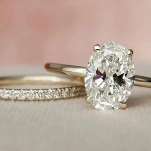 May include: Two gold rings. One ring features a large, oval-cut diamond. The second ring is a thin band with small, sparkling stones. The rings are set against a soft, pink background.