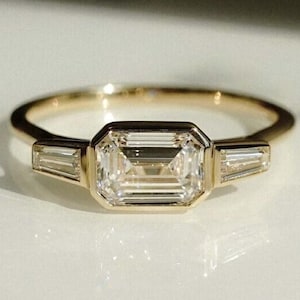May include: A gold ring with a large, rectangular-cut diamond centre stone, with two smaller, baguette-cut diamonds on either side. The ring has a slender band and a modern design, suitable for engagements or special events.