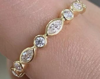 May include: A gold band ring featuring alternating round and marquise-cut clear gemstones. The ring is held between fingers, showcasing the delicate design and sparkle of the gemstones. The ring is a piece of jewelry.