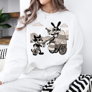 May include: White sweatshirt featuring a black and white cartoon graphic of two characters with guns, one in a vehicle. One character is holding a money bag. The graphic is in a vintage cartoon style.