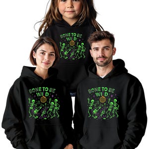May include: Black hooded sweatshirts with the phrase "BONE TO BE WILD" and a neon green skeleton graphic. The graphic includes skeletons dancing around a disco ball. The sweatshirts are worn by three people.