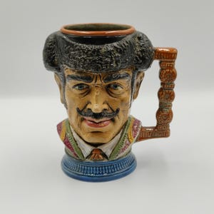 May include: A ceramic character mug shaped like a person wearing a black fur hat, a white collared shirt, and a green and red vest. The handle is brown with a stacked circular pattern. The base is blue with a ribbed texture and a brown rim.