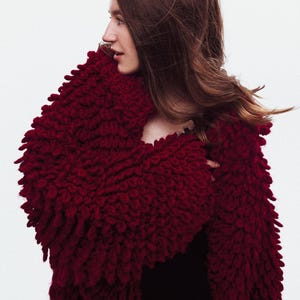 May include: A deep red, textured knit shawl with a fringed edge. The shawl is wrapped around the shoulders, creating a cozy and stylish look. The garment is made of a thick, warm material.