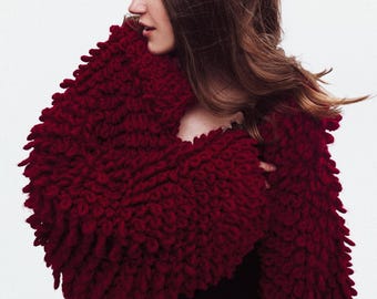 Puede incluir: Un chal de punto texturizado de color rojo intenso con un borde con flecos. El chal está envuelto alrededor de los hombros, creando un look acogedor y elegante. La prenda está hecha de un material grueso y cálido.