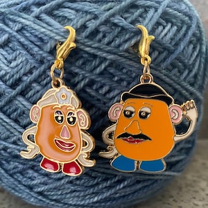 May include: Two gold-coloured metal yarn bowl markers featuring characters from the Toy Story film. One marker features a cartoon image of Mrs. Potato Head with a white hat and red shoes. The other marker features a cartoon image of Mr. Potato Head with a black hat and blue trousers.