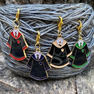 May include: Four gold-colored stitch markers shaped like Hogwarts house robes. The robes are black with different colored accents: red, cream, green, and blue.