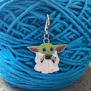 May include: A blue yarn ball with a silver and green metal charm of a character from the Star Wars franchise. The character is wearing a white robe and holding a small cup.