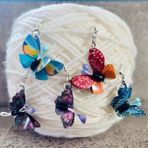 Puede incluir: Cuatro pendientes de mariposa coloridos con ganchos plateados. Las mariposas están hechas de papel y tienen diferentes patrones y colores, incluyendo azul, rojo, rosa y negro.