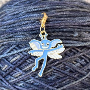 May include: A blue and gold metal stitch marker featuring a cartoon fairy with wings. The fairy has a smiling face and is holding its arms out.