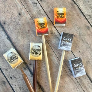 May include: Miniature book-themed party decorations. The image shows several small, colorful book covers attached to wooden sticks. The titles include "Iron Flame", "Fourth Wing", and "Onyx Storm". The books have a fantasy theme.