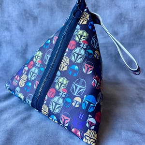May include: A triangular zippered pouch with a white strap. The pouch is made of black fabric with a repeating pattern of colorful Mandalorian helmets.