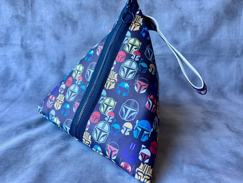 May include: A triangular zippered pouch with a white strap. The pouch is made of black fabric with a repeating pattern of colorful Mandalorian helmets.