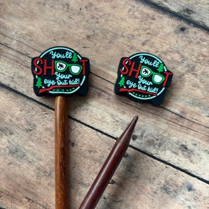 May include: Two wooden knitting needle toppers with the text "You'll shoot your eye out kid!" in red and white on a black background. The toppers have a Christmas tree and glasses graphic. The needles are brown.
