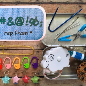 Puede incluir: Una lata de metal con un diseño de tejido de punto azul y blanco en la tapa. La tapa dice "*#&@!%$"; rep from*". La lata contiene una variedad de artículos de costura y tejido, incluyendo un par de tijeras, agujas, un gancho de ganchillo, una cinta métrica, marcadores de puntadas y un tazón de hilo.