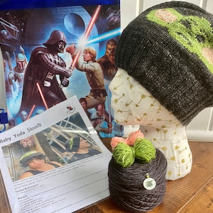 May include: A knitting kit for a Baby Yoda Slouch hat, featuring a black and green yarn, a pattern, and a bag with a Star Wars image. The pattern includes instructions for the hat and a list of materials needed.