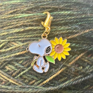 Op de afbeelding: Een bedel met een stripfiguur die een zonnebloem vasthoudt. De bedel heeft een goudkleurige sluiting en een wit, blauw en geel kleurenschema. De zonnebloem heeft gele bloemblaadjes en een bruin hart. De bedel is waarschijnlijk voor een armband of ketting.