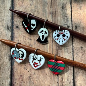 May include: Set of six heart-shaped stitch markers with horror movie character designs. Each marker features a different character, including Jason Voorhees, Ghostface, and others, in black, white, red, and green. They are attached to wooden knitting needles.