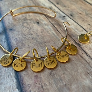 May include: A gold bangle bracelet with seven gold charms. The charms are engraved with the words "Beg", "Knit", "Purl", "SSK", "K2tog", "YO", and "End".