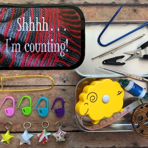 May include: A metal tin with a colourful knitted fabric design on the lid. The lid says "Shhh... I'm counting!" The tin is open and contains a variety of knitting supplies, including stitch markers, a tape measure, a pair of scissors, and a needle.