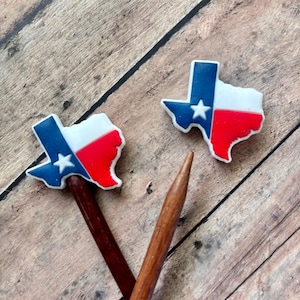 May include: Two Texas-shaped charms with the state flag design, featuring blue, white, and red colors, and a white star. The charms are attached to dark brown wooden sticks, set against a wooden background.