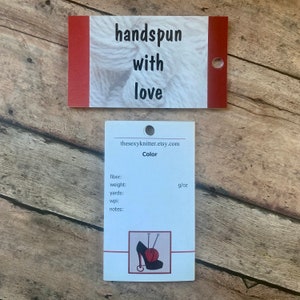 May include: Two white and red tags on a wooden surface. The top tag reads "handspun with love." The bottom tag has the text "thesexyknitter.etsy.com" and "Color." It also includes fields for fiber, weight, yards, wpi, and notes, with a g/oz measurement.