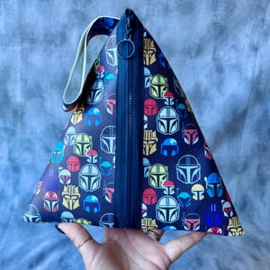 May include: A triangular zippered pouch with a black background and a repeating pattern of colorful Mandalorian helmets. The pouch has a black zipper and a white strap.