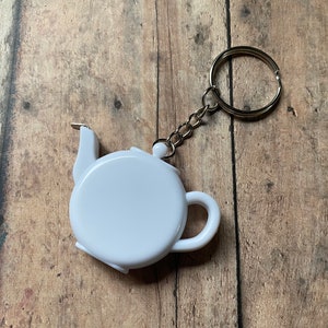 May include: White teapot-shaped keychain with a retractable tape measure. The keychain has a silver metal ring and chain.