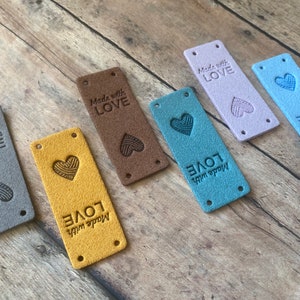 Pode incluir: Seis etiquetas de tecido de diferentes cores com o texto "Made with Love" e um coração com fio dentro. As etiquetas são marrom, amarelo, azul, azul claro, roxo e cinza.