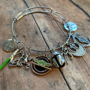 May include: A silver charm bracelet with multiple charms, including a green lightsaber, a frog, a Mandalorian helmet, a Star Wars logo, and a charm that says "This is the way".