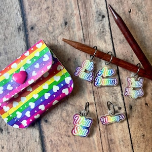May include: A small rainbow-striped pouch with white hearts and a pink heart snap closure, accompanied by five metal stitch markers featuring rainbow-coloured text that reads Oh Fuck, Aw Damn, Crud Nuggets, Well Shit, and Whoops on a wooden needle.
