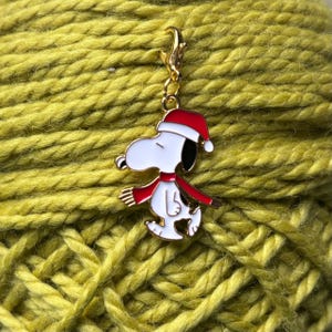 May include: A gold metal charm featuring a cartoon character wearing a red Santa hat and a red scarf. The character is white with black eyes and a black nose. The charm is attached to a gold metal chain.