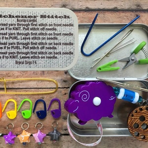 May include: A metal tin with a purple sheep-shaped tape measure, a wooden yarn bowl, and a variety of knitting needles, stitch markers, and other tools. The tin has a label that reads "Kitchener Stitch" and includes instructions for the stitch.