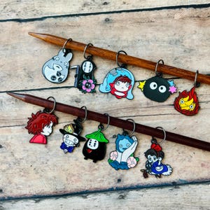 May include: A collection of colorful enamel stitch markers featuring characters from animated films. The markers are attached to wooden knitting needles. The characters include a gray Totoro, a black No-Face, and a red-haired girl.