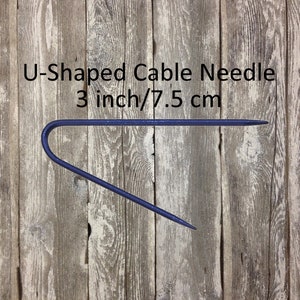 May include: A blue U-shaped cable needle, 3 inches or 7.5 cm long, against a wood background.
