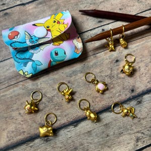 May include: A collection of gold-colored stitch markers with various character designs, including a Pikachu, and a small pouch with a button closure. The pouch features a colorful print of cartoon characters on a wooden surface.