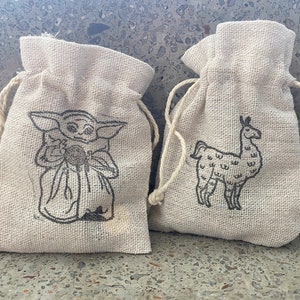 May include: Two small, beige burlap drawstring bags with black embroidered designs. One bag features a cartoon image of a character from the Star Wars franchise, "The Mandalorian", known as "Baby Yoda". The other bag features a cartoon image of a llama.