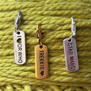 Könnte beinhalten: Drei silberne und goldene Metall-Maschenmarkierer mit verschiedenen Designs. Der erste Marker ist silber und sagt "I ♥ DR WHO". Der zweite Marker ist golden und sagt "TREKKIE" mit einem Herzen. Der dritte Marker ist silber und sagt "STAR WARS".