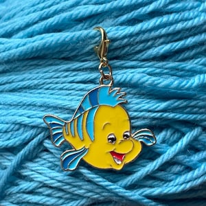 Op de afbeelding: Een gele en blauwe emaille bedel in de vorm van een cartoonvis, met een goudkleurige sluiting. De bedel is geplaatst tegen een achtergrond van blauw garen, wat duidt op een knutselthema.