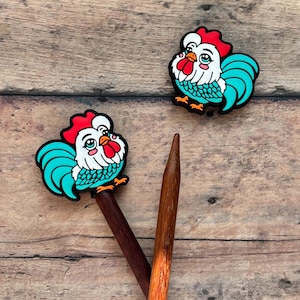 Puede incluir: Dos adornos para agujas de tejer con forma de gallo, con crestas rojas, caras blancas y plumas de cola turquesas. Los adornos están sobre agujas de tejer de madera, sobre un fondo de madera.