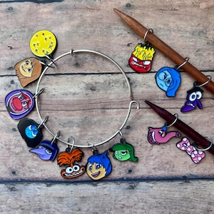 May include: A silver bangle bracelet with colorful enamel charms featuring characters from the movie Inside Out. The charms include Joy, Sadness, Anger, Fear, Disgust, Bing Bong, and a star. The bracelet is also shown with two wooden knitting needles with additional charms.