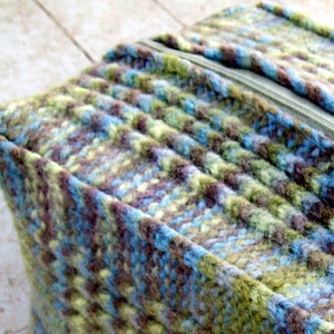 May include: A close-up of a zipper on a knitted fabric bag. The bag is made of a blue, green, and brown yarn with a subtle pattern. The zipper is silver and has a pull tab.