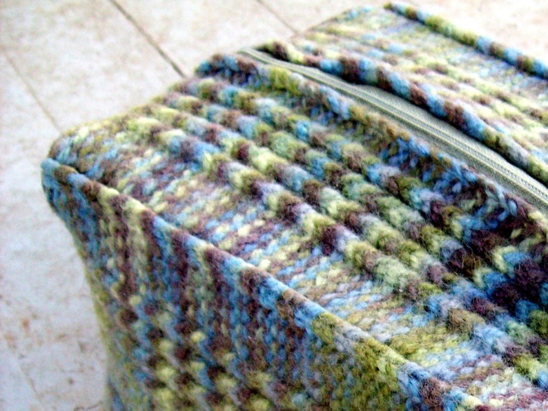 May include: A close-up of a zipper on a knitted fabric bag. The bag is made of a blue, green, and brown yarn with a subtle pattern. The zipper is silver and has a pull tab.