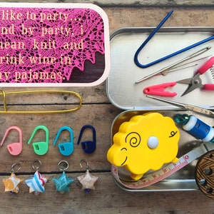 May include: A metal tin filled with knitting supplies, including a tape measure shaped like a sheep, stitch markers, a pair of scissors, and a needle threader. The tin is open and the supplies are visible. The tin is sitting on a wooden surface. A quote about knitting and drinking wine is visible in the background.