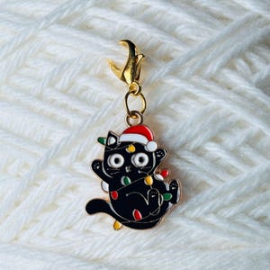 Puede incluir: Un gato negro con un gorro de Papá Noel está enredado en luces de Navidad. El gato es un amuleto con un lazo dorado para sujetarlo a un proyecto.