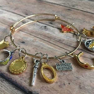May include: A gold bangle bracelet with charms, including a sword, a shield, a star, a crown, and a Wonder Woman logo charm. The charms are all gold and silver.