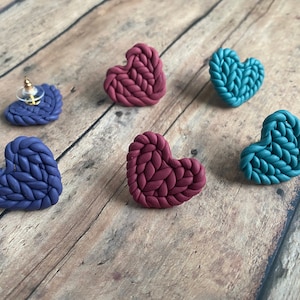 May include: A set of three pairs of heart-shaped earrings. The earrings are made of polymer clay and have a knitted texture. The colors are teal, burgundy, and navy blue.