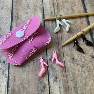 May include: Pink coin purse with a Barbie pattern and a flap closure. Four pairs of miniature high heel shoes in pink, white and black are hanging from key rings.
