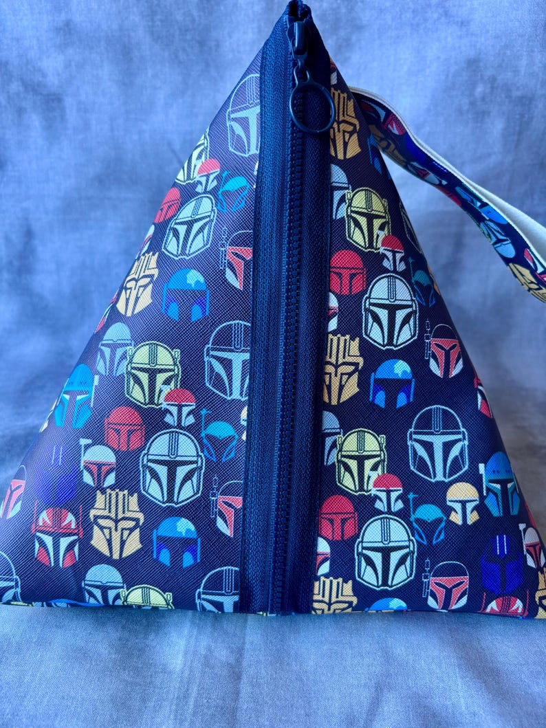 May include: A black zippered pouch with a repeating pattern of colorful Mandalorian helmets. The pouch has a black zipper and a white strap.