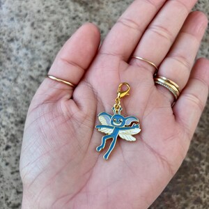 May include: A gold-coloured charm featuring a blue and white cartoon fairy design. The fairy has large eyes, a small mouth, and outstretched wings.
